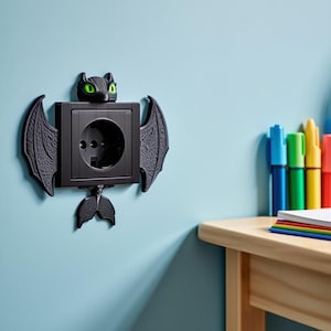 May include: A black wall outlet cover shaped like a dragon, with green eyes, wings, and a tail. The outlet cover is mounted on a light blue wall. A wooden table and colorful markers are in the background.