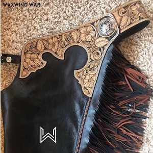 Puede incluir: Primer plano de unos chaps de cuero negro con intrincados dise&ntilde;os florales en cuero marr&oacute;n claro. Los chaps presentan flecos marrones y un concho plateado. La parte superior tiene un cintur&oacute;n con una hebilla plateada. La marca "WAXWING WARI" es visible.