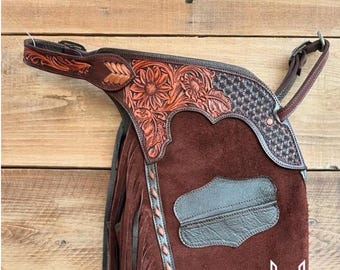 May include: Close-up of brown leather chaps with floral tooling and fringe. The chaps feature a sunflower design and a dark brown leather patch. The top portion has a patterned design. The image shows the brand name "WAXWING WARI".