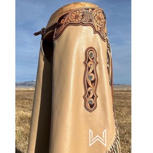 Op de afbeelding: Tan lederen chaps met ingewikkelde bloemmotieven langs de boven- en zijkant. De chaps hebben decoratieve klinknagels en een wit logo aan de onderkant. De achtergrond toont een veld en een blauwe lucht.