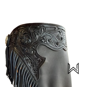 Puede incluir: Chaps de cuero negro con un diseño floral y flecos. Los chaps tienen un concho plateado decorativo en la parte delantera.