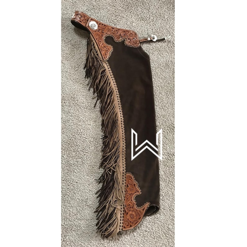 Puede incluir: Chaps de cuero marr&oacute;n con flecos y tallado intrincado. Los chaps tienen una hebilla plateada y un panel de gamuza marr&oacute;n.