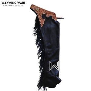 以下が含まれることがあります： 黒い革のチャップス。装飾的なツール加工された革製のヨークが付いています。ヨークは茶色で、複雑な花の模様と小さな正方形の装飾が施されています。チャップスの側面には長い黒いフリンジがあり、下部には白いロゴがあります。「WAXWING WARI CRAFTING LEGACY」の文字が上部にあります。
