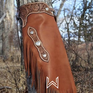 Op de afbeelding: Bruine leren chaps met franjes en zilveren hartvormige conchos. De chaps zijn versierd met ingewikkelde bloemmotieven.
