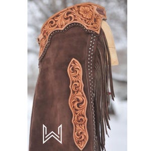May include: Brown suede chaps with intricate floral tooling and fringe. The chaps have a decorative 'W' logo on the front.