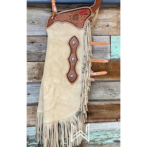 May include: A pair of tan leather chaps with fringe and a tooled leather design. The chaps have three silver conchos and a red and brown tooled leather design on the top.
