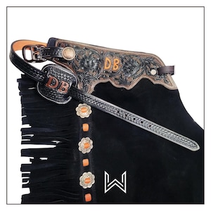 Könnte beinhalten: Schwarze Lederchaps mit Fransen, einem geprägten Lederdesign und einem schwarzen Ledergürtel mit Schnalle. Die Chaps haben orangefarbene Nähte und Metallakzente. Der Gürtel hat die Initialen "DB" geprägt.