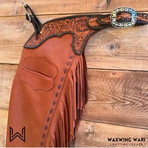 May include: Brown leather chaps with detailed floral tooling and fringe. The chaps have a decorative belt with a silver buckle. The image also shows the Waxwing Wari logo.