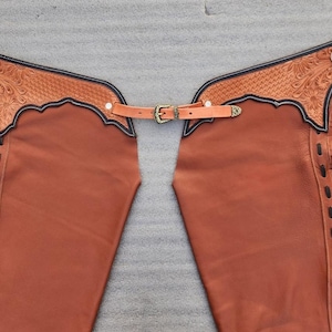 Pu&ograve; includere: Chaps in pelle marrone con decorazioni floreali sul giogo e frange sui lati. Gli chaps hanno una cintura con fibbia in ottone e sono progettati per l'equitazione. La pelle &egrave; di un caldo colore marrone-rossiccio.