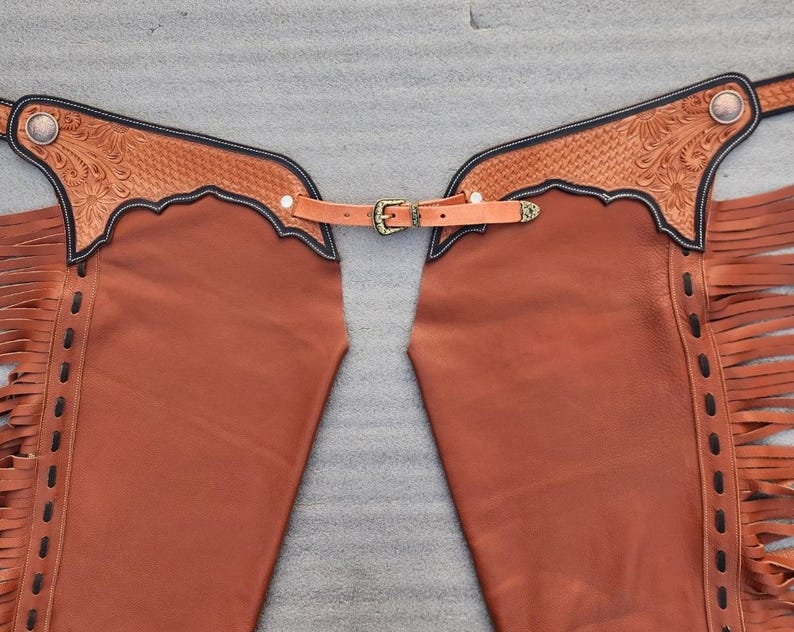 Pu&ograve; includere: Chaps in pelle marrone con decorazioni floreali sul giogo e frange sui lati. Gli chaps hanno una cintura con fibbia in ottone e sono progettati per l'equitazione. La pelle &egrave; di un caldo colore marrone-rossiccio.