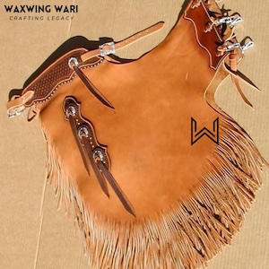 Op de afbeelding: Bruine leren chaps met zilverkleurige hardware en franjes langs de onderkant. De chaps hebben een riem- en gespsluiting. De tekst "WAXWING WARI CRAFTING LEGACY" staat linksboven.