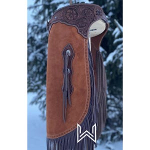May include: Brown leather chaps with intricate floral tooling and fringe. The chaps have a suede front panel and a leather back panel. The fringe is brown and the tooling is silver.