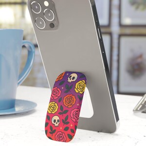 May include: A smartphone is propped up by a colorful phone grip. The grip features a pattern of roses, skulls, and leaves in shades of red, yellow, and purple. The phone is gray, and a blue mug is in the background.