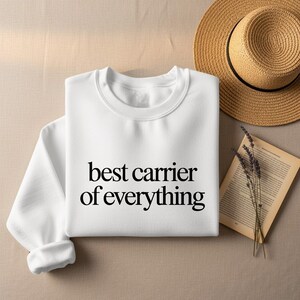 White crewneck sweatshirt with "best carrier of everything" text, next to a hat and book.
