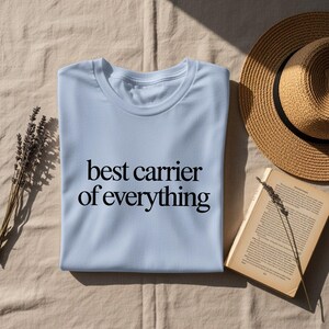 Light blue t-shirt with "best carrier of everything" text, next to a straw hat and book.