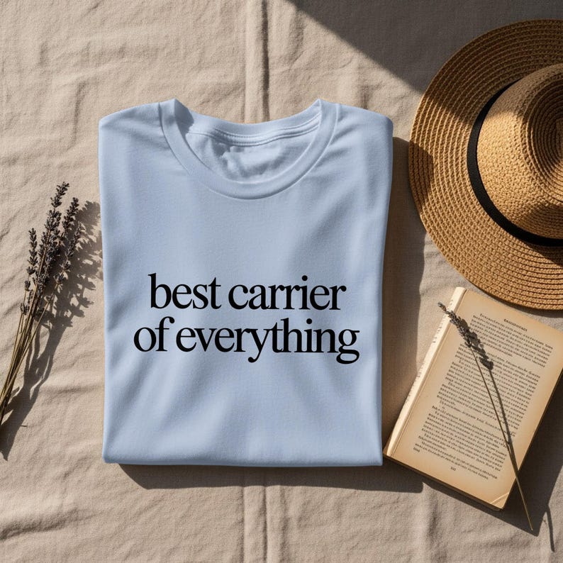 Light blue t-shirt with "best carrier of everything" text, next to a straw hat and book.