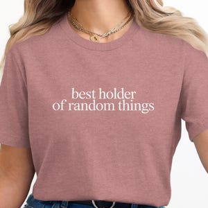 May include: A dusty rose-colored t-shirt with the phrase "best holder of random things" printed in white. The shirt is a crew neck style and made of a soft material. The overall theme is casual wear.