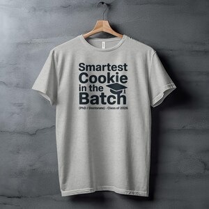 May include: A light gray t-shirt with the text "Smartest Cookie in the Batch" in dark blue, along with a graduation cap graphic. Below the text is "(PhD / Doctorate) - Class of 2026". The shirt is hanging on a wooden hanger.