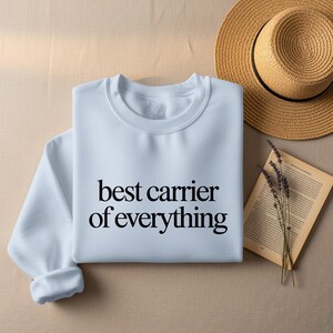 White sweatshirt with "best carrier of everything" text, hat and book.