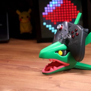 Pode incluir: Um suporte de controlador verde e preto em forma de Rayquaza, um Pokémon semelhante a um dragão. O suporte tem uma boca aberta com detalhes rosa e um olho amarelo. Um controlador preto repousa no topo. A palavra "FUTURE" está escrita no controlador.
