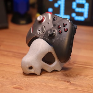 May include: A gray and black video game controller with red buttons is resting in a gray 3D printed stand. The controller has the words "No. Filter" written on it.