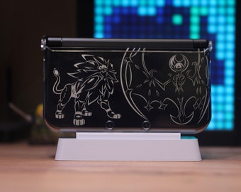 May include: A black Nintendo 3DS handheld gaming console with a white stand. The console has a custom design featuring a black and white silhouette of a lion and a bat.