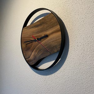 May include: A round wooden wall clock with a black metal frame. The clock face is made of a dark wood with a natural grain pattern. The clock hands are black and red.