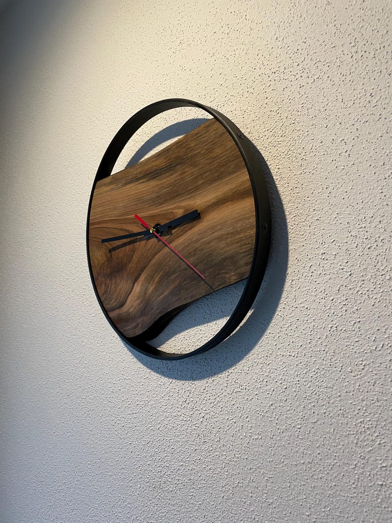 May include: A round wooden wall clock with a black metal frame. The clock face is made of a dark wood with a natural grain pattern. The clock hands are black and red.