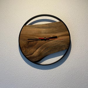 May include: A round wooden clock with a black metal frame. The clock face is made of a natural wood slice with a unique grain pattern. The clock hands are black and red.