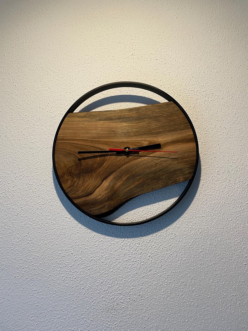 May include: A round wooden clock with a black metal frame. The clock face is made of a natural wood slice with a unique grain pattern. The clock hands are black and red.