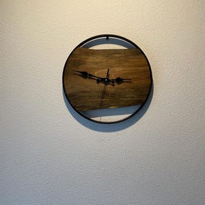 May include: A round wooden clock with black hands and a black metal frame. The clock is mounted on a white wall.