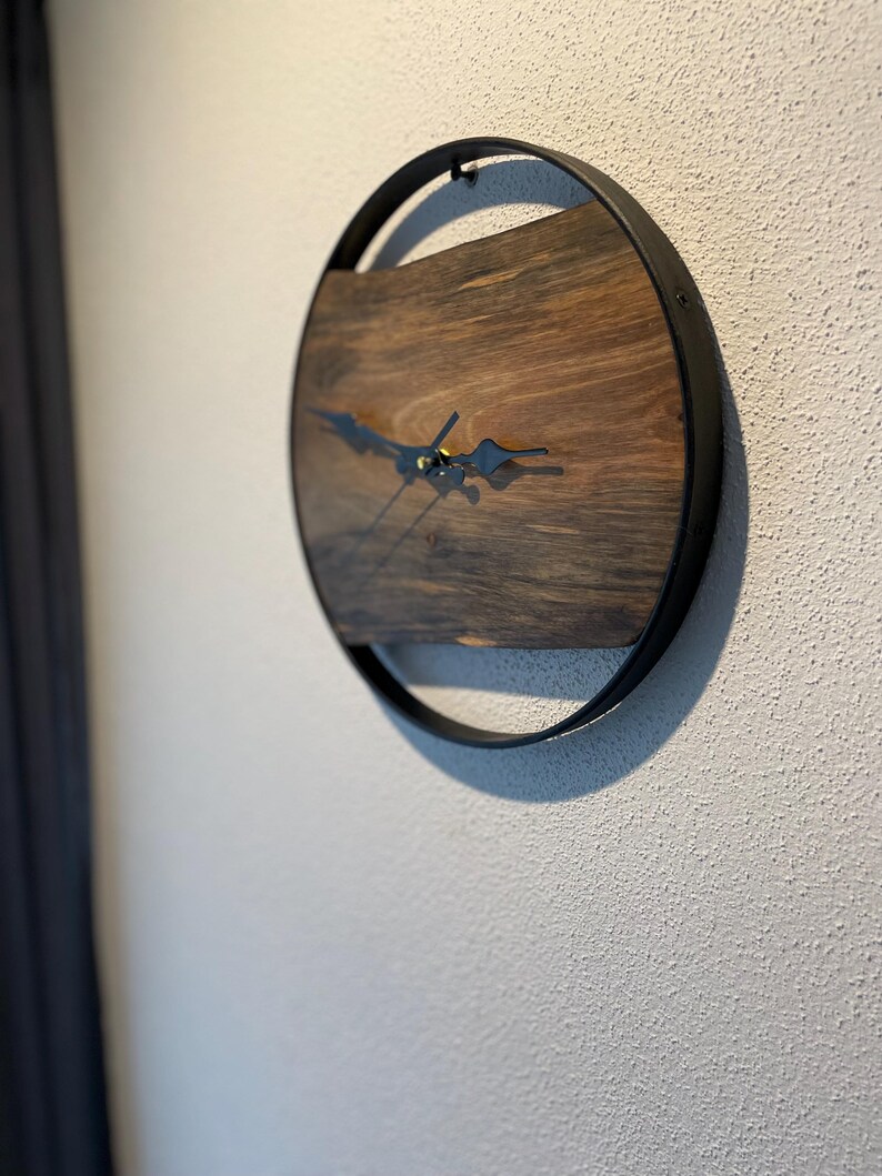 May include: A round wooden clock with black metal hands and a black metal frame. The clock face is made of dark wood with a natural grain pattern.