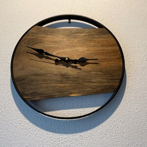 May include: A round wooden clock with black metal hands and a black metal frame. The clock face is made of a dark wood with a natural grain pattern.