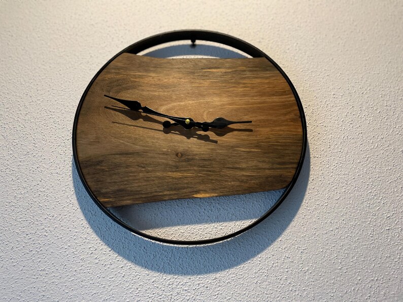 May include: A round wooden clock with black metal hands and a black metal frame. The clock face is made of a dark wood with a natural grain pattern.