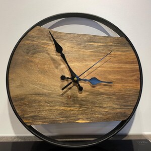 May include: A round wooden clock with black hands and a black metal frame. The clock face is made of a dark wood with a natural grain pattern.