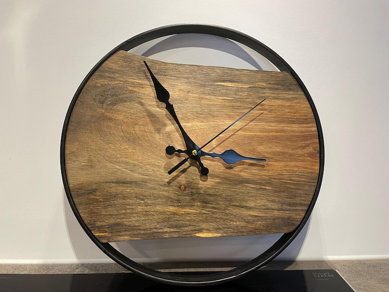 May include: A round wooden clock with black hands and a black metal frame. The clock face is made of a dark wood with a natural grain pattern.