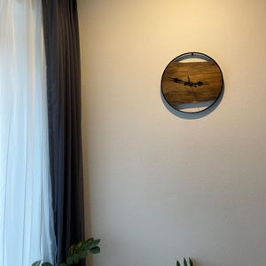 May include: A round wooden clock with black hands and a black metal frame. The clock is hanging on a white wall.