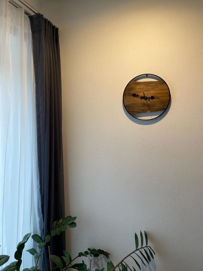 May include: A round wooden clock with black hands and a black metal frame. The clock is hanging on a white wall.