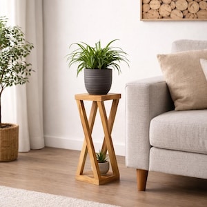 Könnte beinhalten: Ein hölzerner Pflanzenständer mit X-förmigem Fuß trägt einen dunkelgrauen Topf mit einer grünen Pflanze. Darunter befindet sich eine kleinere Pflanze in einem hellen Topf. Der Ständer steht auf einem Hartholzboden in der Nähe eines hellen Sofas.