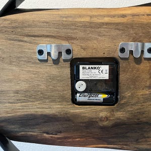 May include: The back of a wooden clock with a black metal frame. The clock mechanism is visible, along with a battery labeled "Energizer Industrial". The clock has two metal hanging brackets on the back.