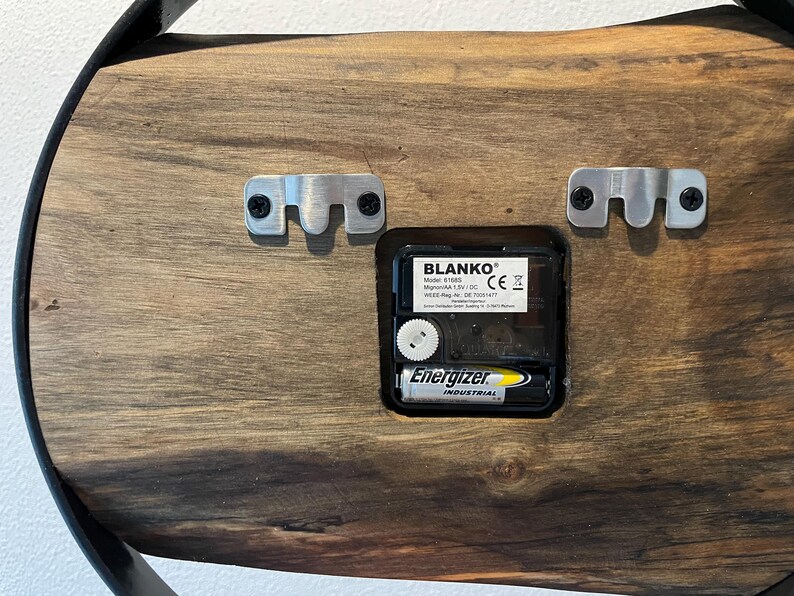 May include: The back of a wooden clock with a black metal frame. The clock mechanism is visible, along with a battery labeled "Energizer Industrial". The clock has two metal hanging brackets on the back.