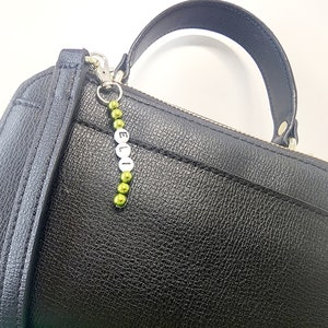 May include: A black leather handbag with a silver charm that says "ELLI" in white letters. The charm is attached to a green beaded chain.