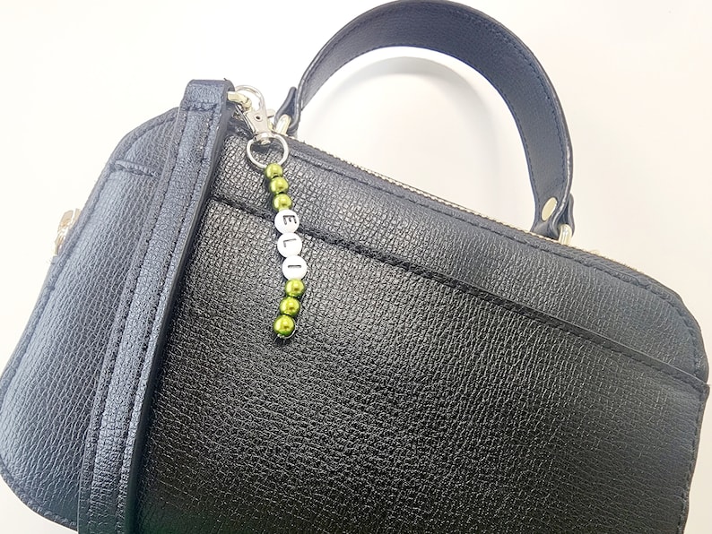 May include: A black leather handbag with a silver charm that says "ELLI" in white letters. The charm is attached to a green beaded chain.