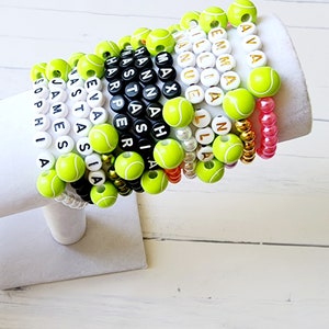 May include: A stack of colorful bracelets with tennis ball beads and names spelled out in black letters. The names include Ava, Emma, Malia,  Manuela, Lila, Nash,  Hastna, Harper, Eva,  Nassia, James, and Sophia.