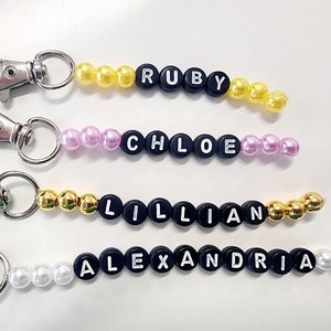 May include: Four keychains with different names spelled out in black beads with white letters. Each keychain has a silver key ring and a different color of beads: yellow, pink, gold, and white.