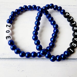 May include: Two beaded bracelets with the names "ZOE" and "ISABELLA" spelled out in black letters. The beads are blue and the letters are white on one bracelet and black on the other.