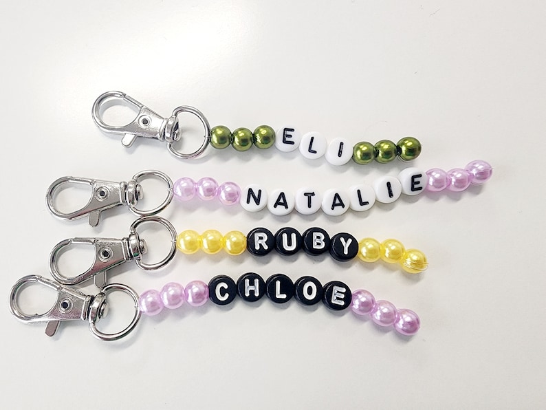 May include: Four personalized keychains with different names spelled out in beads. The names are Ellie, Natalie, Ruby, and Chloe. Each keychain has a silver metal clasp.