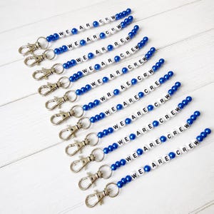 May include: A collection of ten beaded lanyards featuring alternating royal blue spherical beads and white cylindrical beads with black lettering. Each lanyard includes a silver-toned metal lobster clasp at one end, with text spelling out WE ARE CREW.