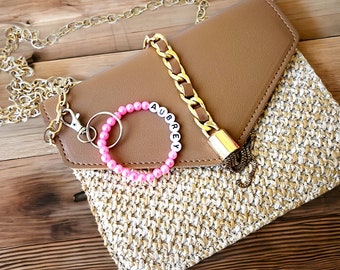 May include: A tan clutch with a gold chain strap and a woven straw bottom. A pink beaded bracelet with the name "AUBREY" is attached to the clutch. A gold chain and lock detail adds a touch of elegance.