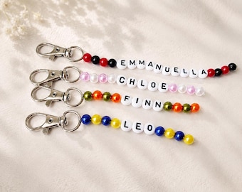 May include: Four keychains with silver-toned lobster clasps and strands of beads. The beads include white letter cubes spelling EMMANUELLA, CHLOE, FINN, and LEO, interspersed with red, black, pink, orange, green, yellow, and blue spherical beads.